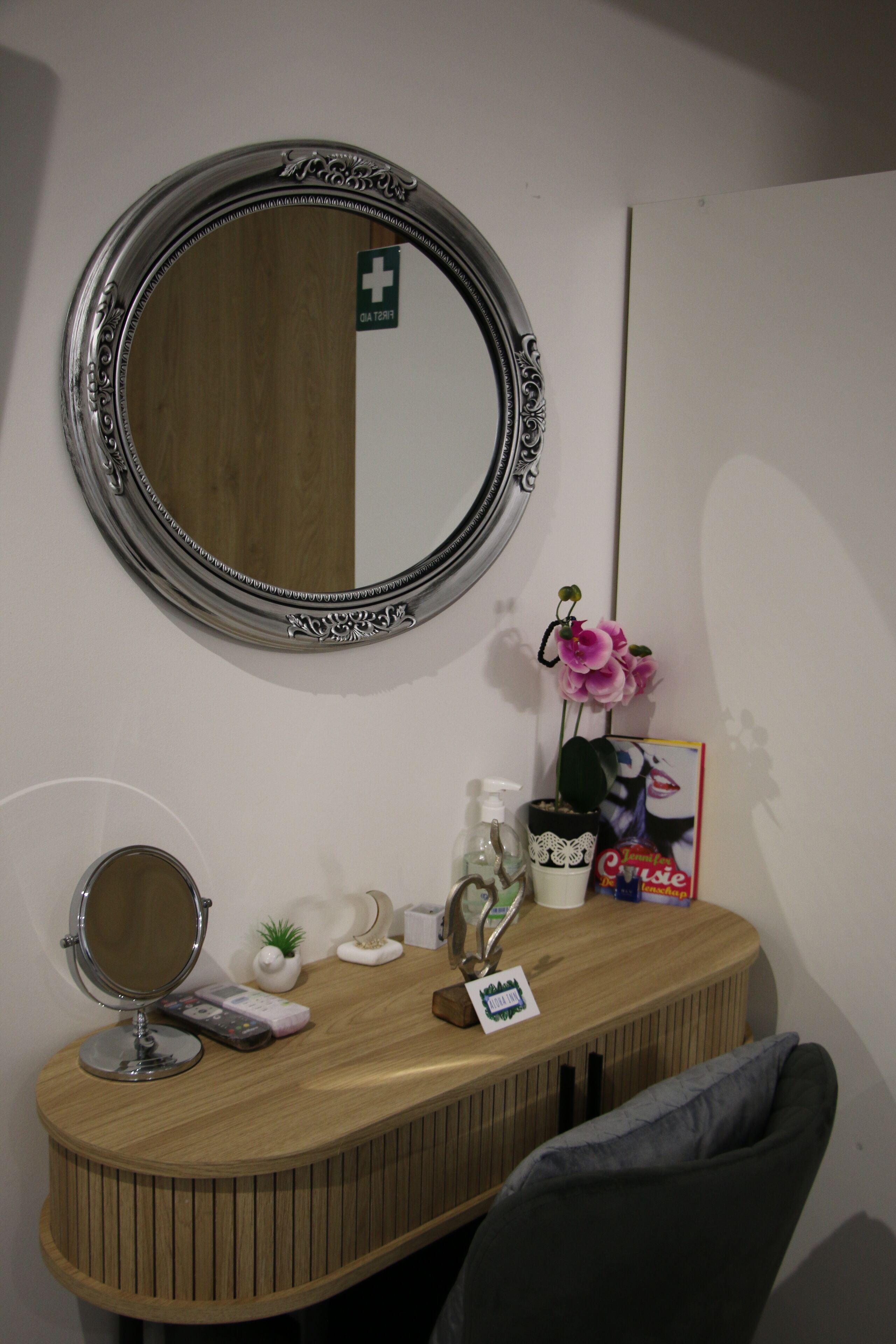 Un coin coiffeuse avec un miroir rond, une petite plante en pot, une bouteille, une boîte de mouchoirs et un magazine, le tout posé sur un comptoir en bois.