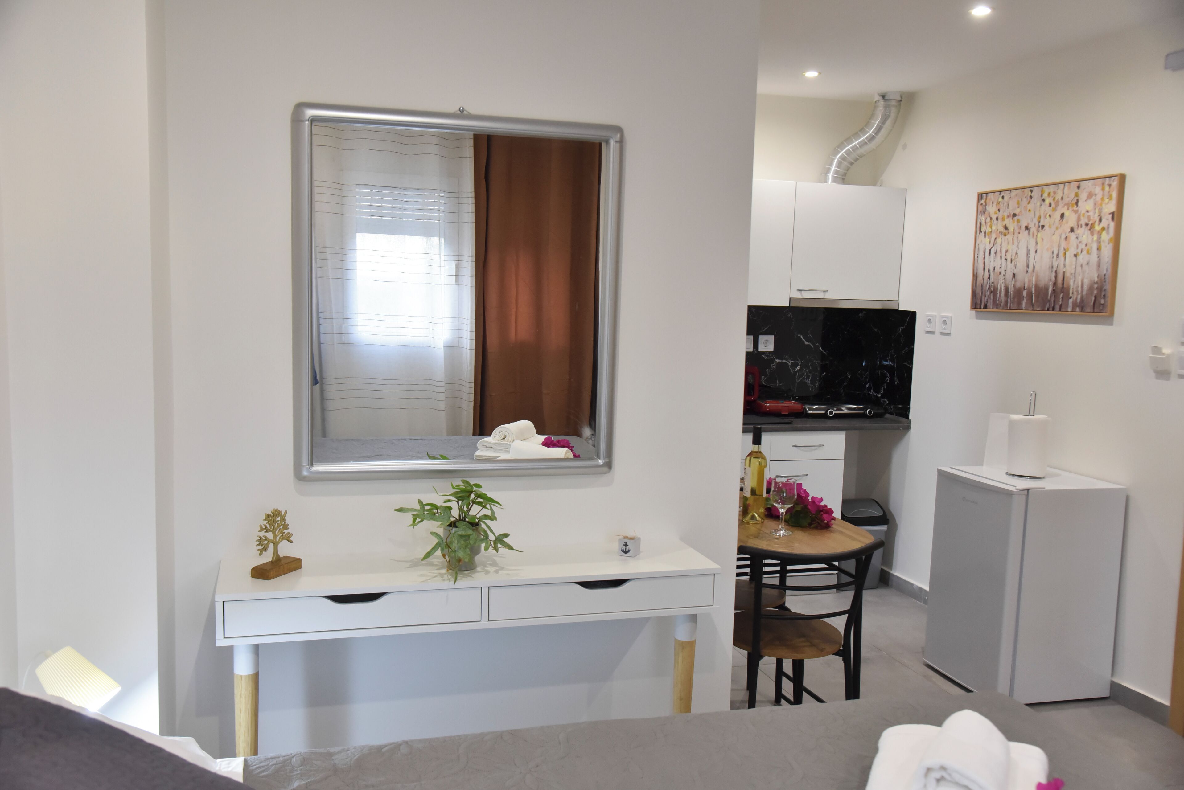 A compact hotel room with a bed, a desk with a mirror, a small dining table, a TV, and a refrigerator.