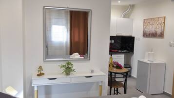 A compact hotel room with a bed, a desk with a mirror, a small dining table, a TV, and a refrigerator.