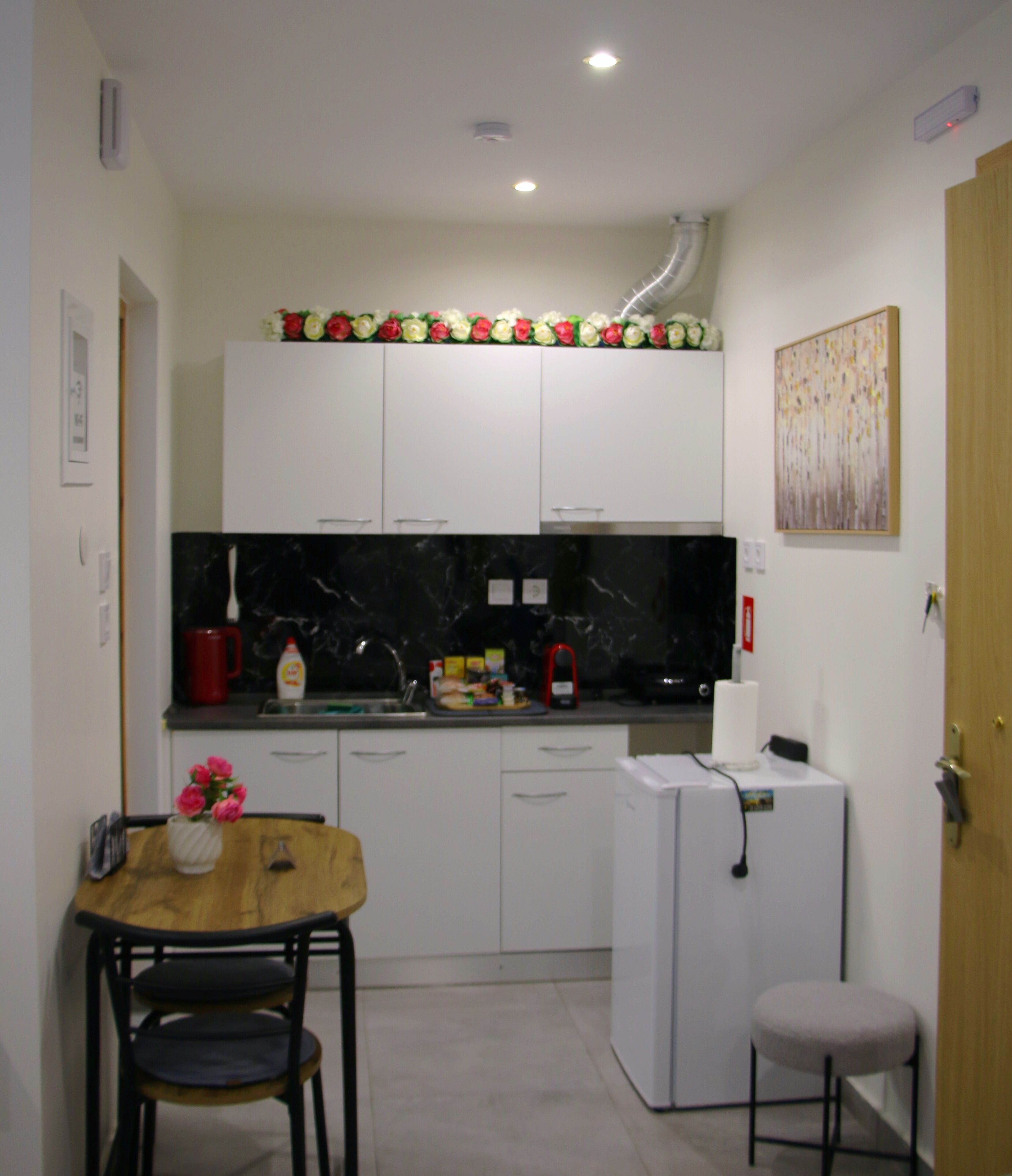 A compact kitchen with white cabinets, a black countertop, a small refrigerator, and a dining area with a wooden table and two chairs.