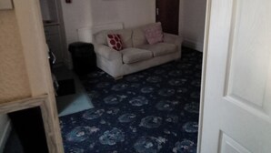 A living room with a patterned carpet, a white sofa with red and pink cushions, and a dark-colored trash bin. - Seaside Escape at Withnell Road (Blackpool)