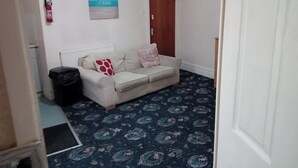 A small, neatly arranged room with a beige sofa, patterned carpet, a fire blanket on the wall, and a door. - Seaside Escape at Withnell Road (Blackpool)
