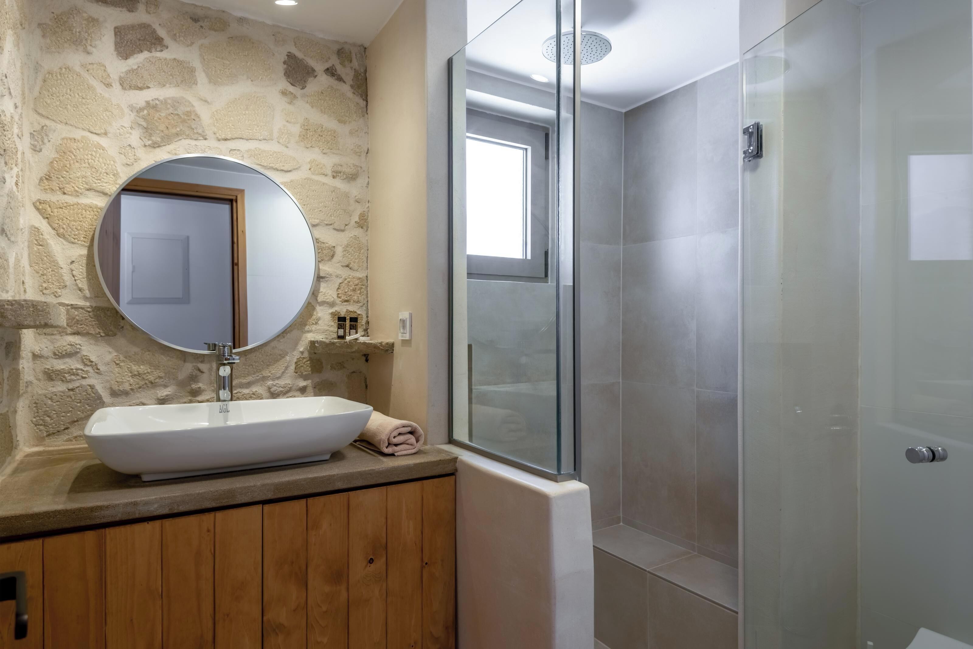 A bathroom with a stone wall, a round mirror, a white sink, and a glass-enclosed shower.
