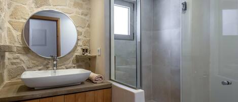 A bathroom with a stone wall, a round mirror, a white sink, and a glass-enclosed shower.