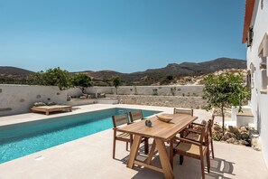 A poolside area with a wooden table and chairs, a lounge chair, and a small tree. - Villa Froudi (Kissamos)