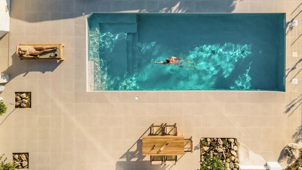 A rectangular swimming pool with a person swimming, surrounded by tiled walls, a wooden table, and a small seating area. - Villa Froudi (Kissamos)