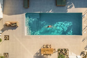 A rectangular swimming pool with a person swimming, surrounded by tiled walls, a wooden table, and a small seating area. - Villa Froudi (Kissamos)