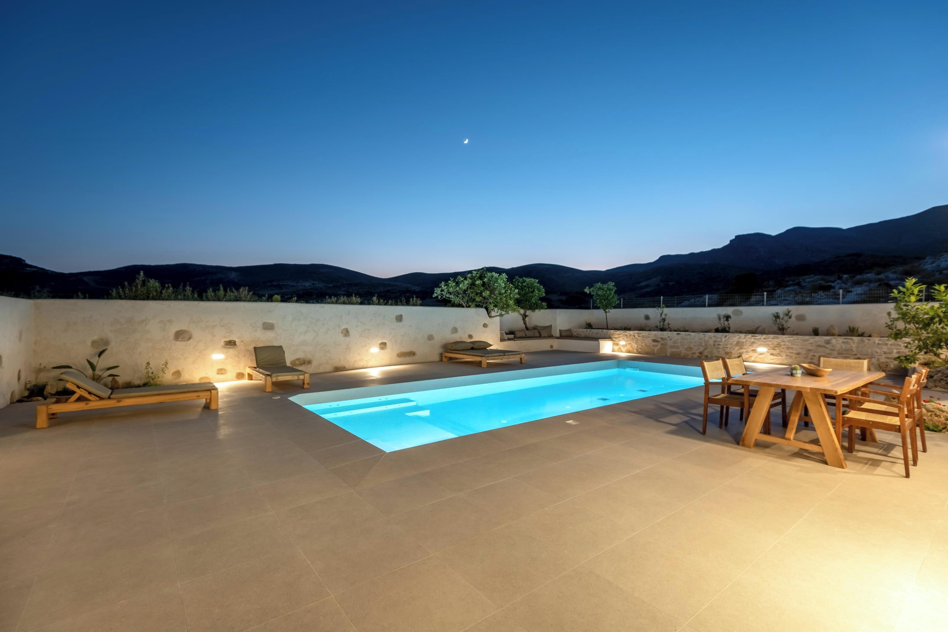 A rooftop pool area with a clear view of the mountains, a wooden table and chairs, and a lounge chair.