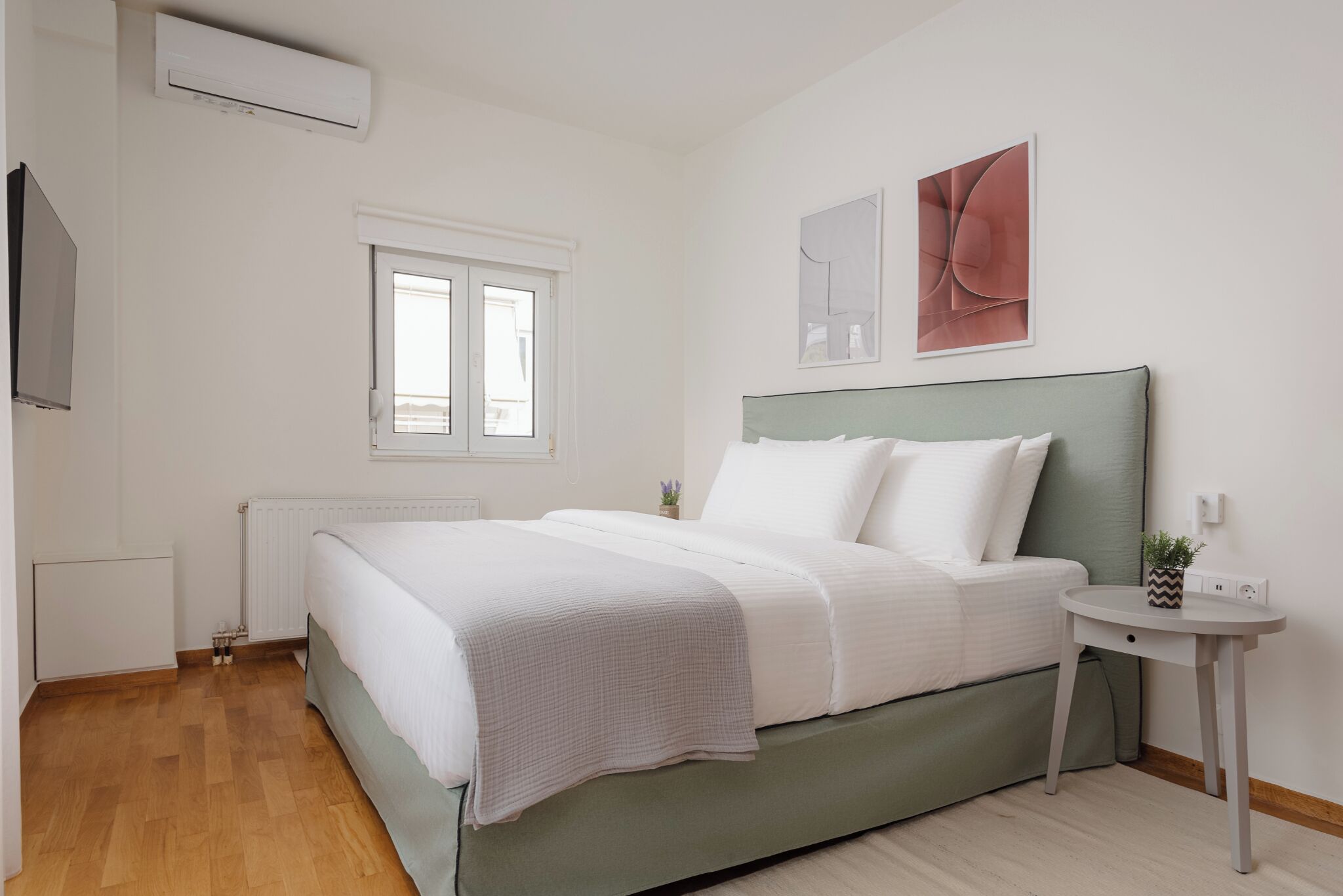 Een moderne slaapkamer met een groen bed, witte beddengoed, een klein nachtkastje met een plant en een raam met zonwering.