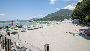 Una spiaggia con file di lettini e ombrelloni, un molo di legno e montagne sullo sfondo.