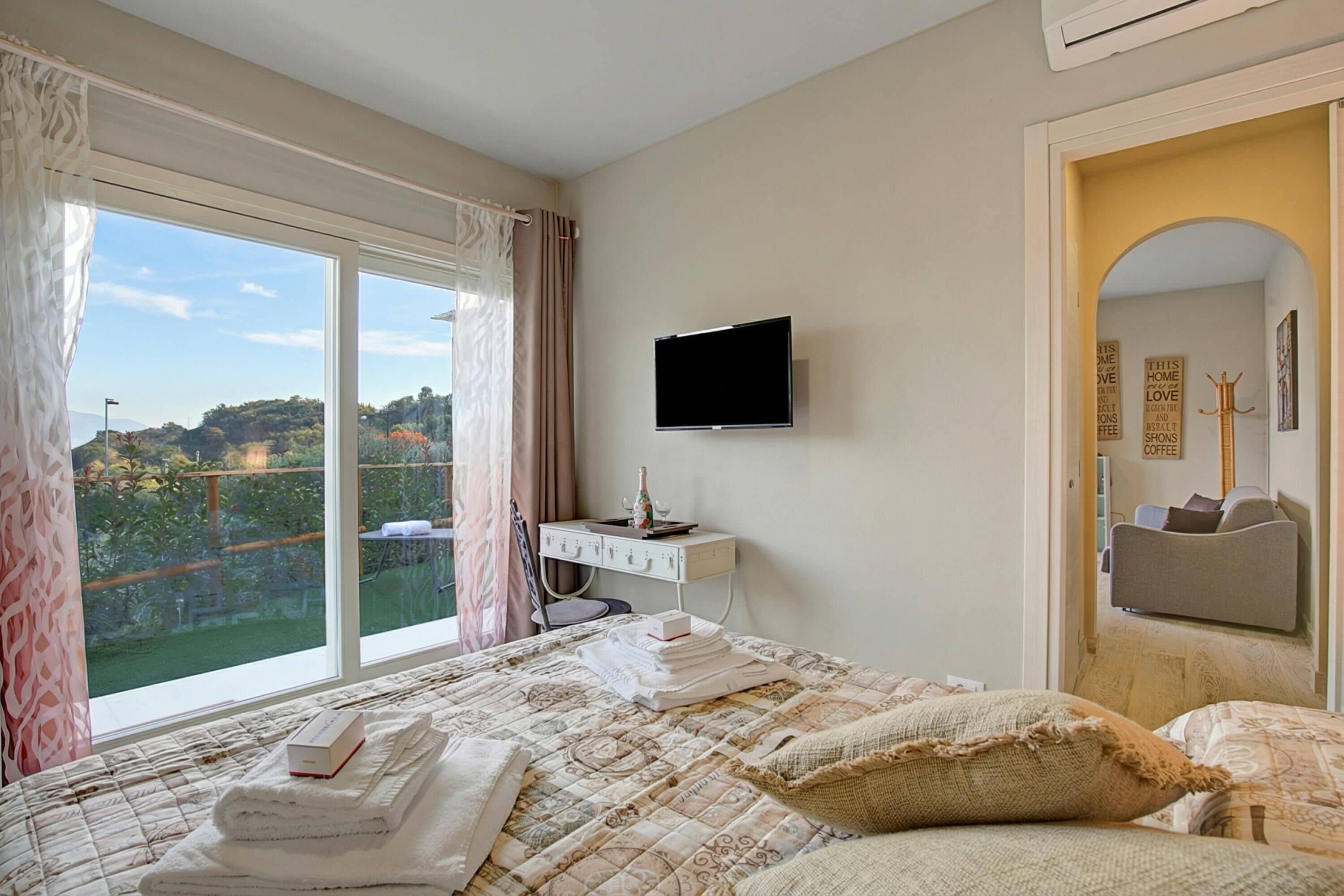Un dormitorio con un ventanal grande, un televisor de pantalla plana, un escritorio con silla y una cama con toallas ordenadas.