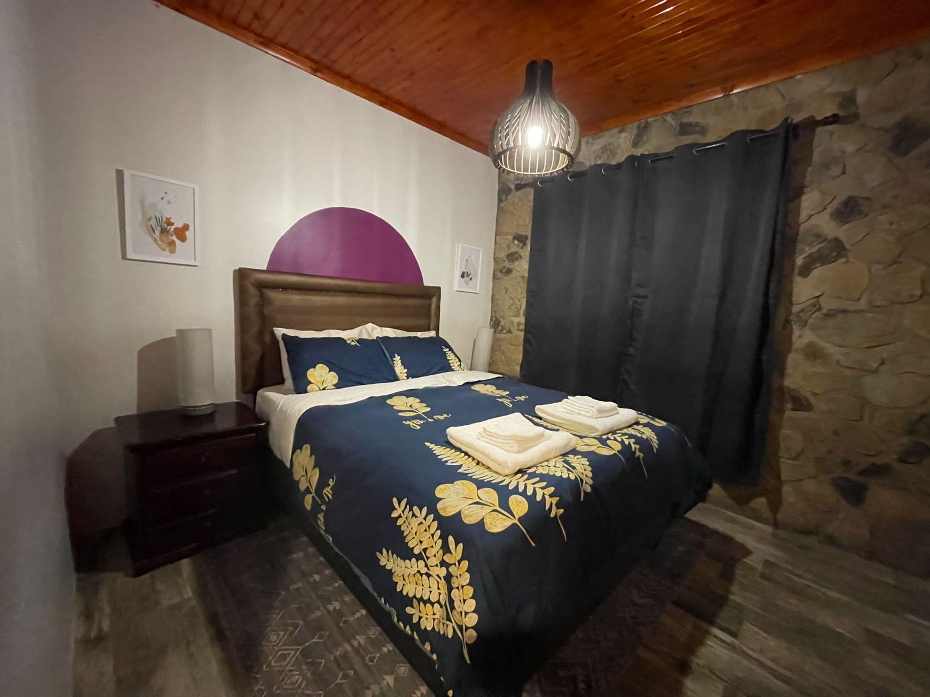 Een slaapkamer met een stenen muur, een houten plafond, een bed met een blauw en gouden bloemenbedekking, een houten nachtkastje en een hangende lamp.