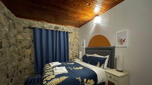 A bedroom with a stone wall, a wooden ceiling, a bed with a blue and gold bedspread, a nightstand with a lamp, and a wall-mounted picture. - Lovely 3 bed House in Juliasdale - 2273 (Nyanga)