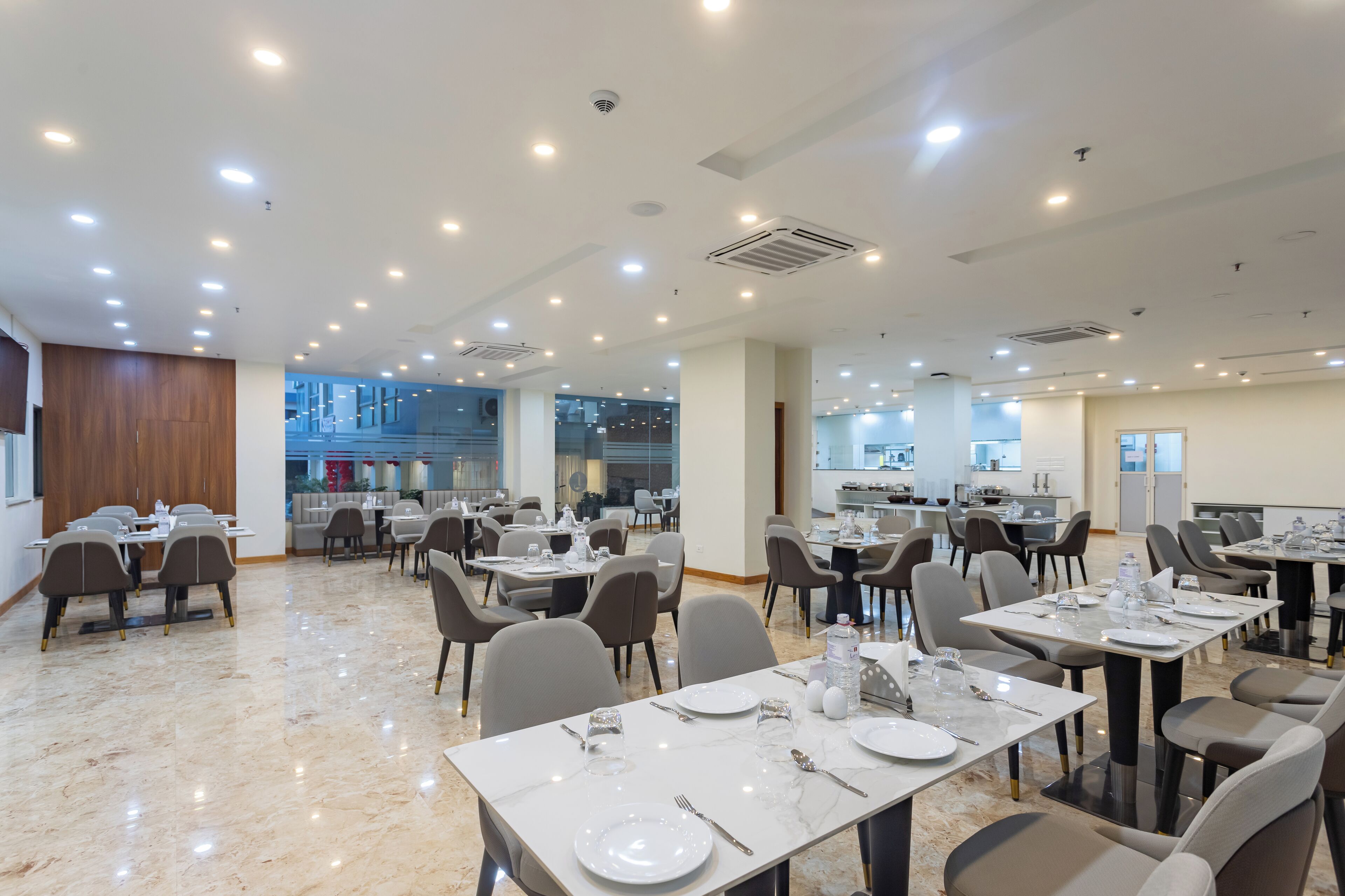 A spacious dining area with multiple tables and chairs, ceiling lights, and a view of the kitchen in the background.