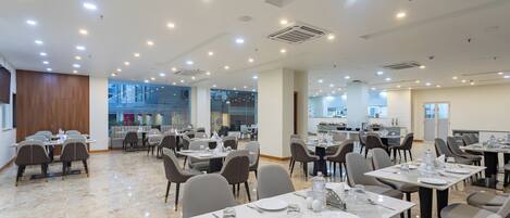 A spacious dining area with multiple tables and chairs, ceiling lights, and a view of the kitchen in the background.