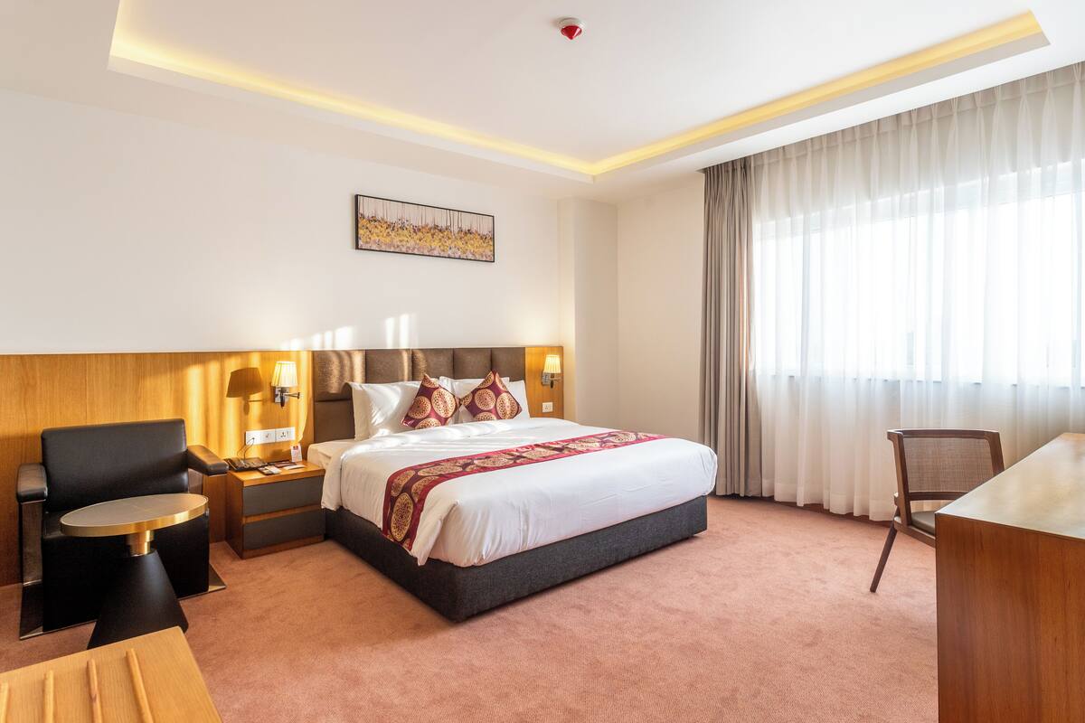 Habitación de hotel con una cama grande, un escritorio, una silla, una mesita de noche y una lámpara de pared.