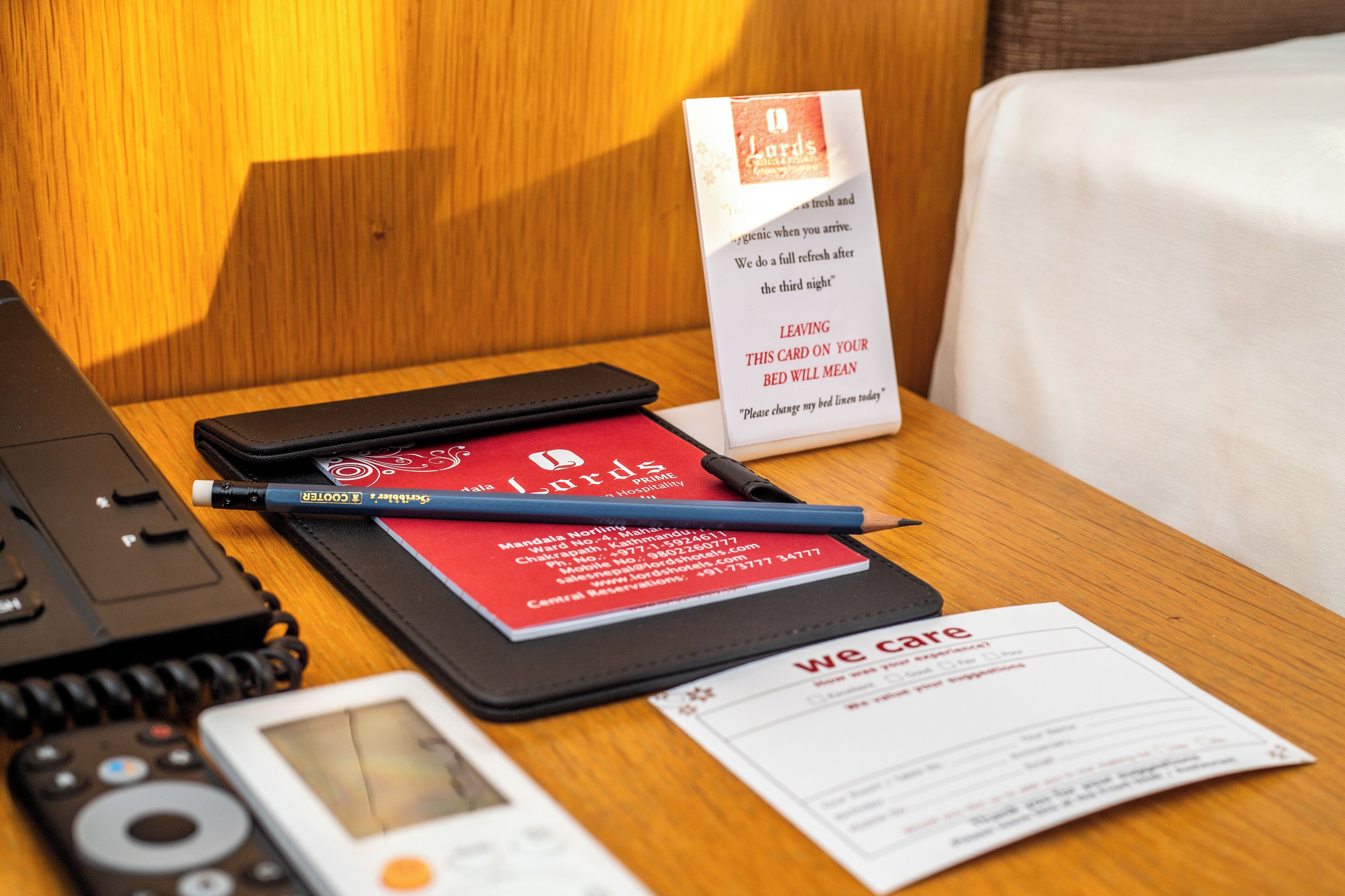 A hotel room with a wooden headboard, a bedside table with a red card, a pen, a smartphone, and a card holder with a card.