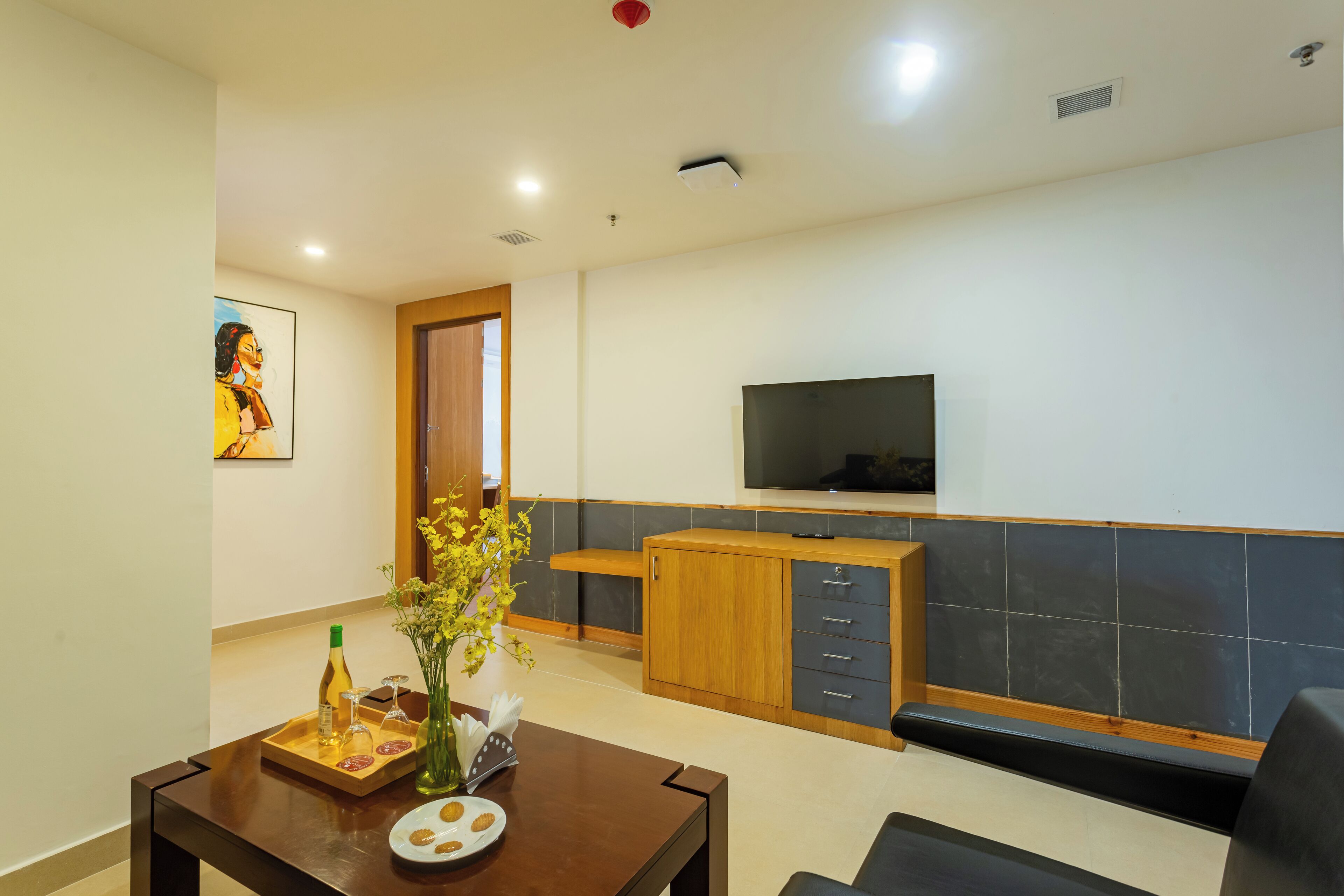 A modern living room with a wooden coffee table, a TV mounted on the wall, and a kitchen area with a wooden cabinet and tiled backsplash.