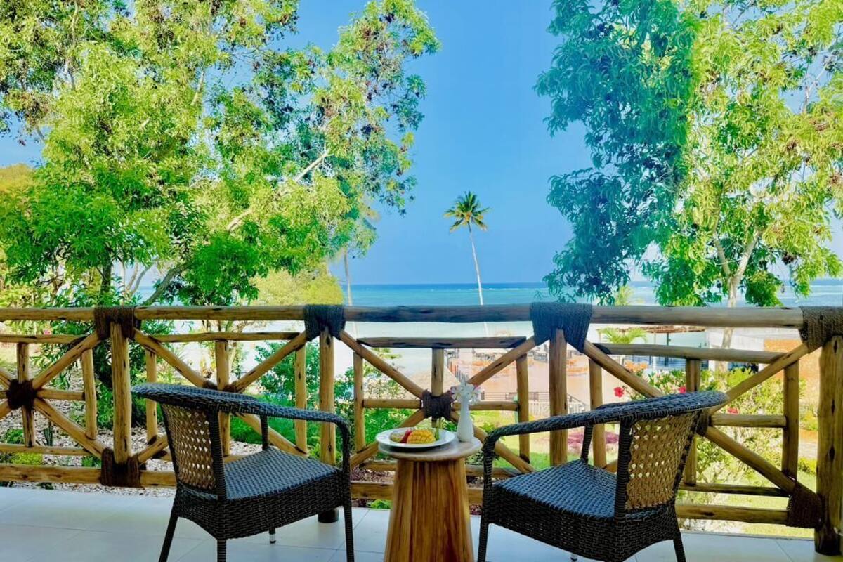 Ein Balkon mit Korbstühlen und einem kleinen runden Tisch, mit Blick auf einen Strand mit Palmen und einem strahlend blauen Himmel.