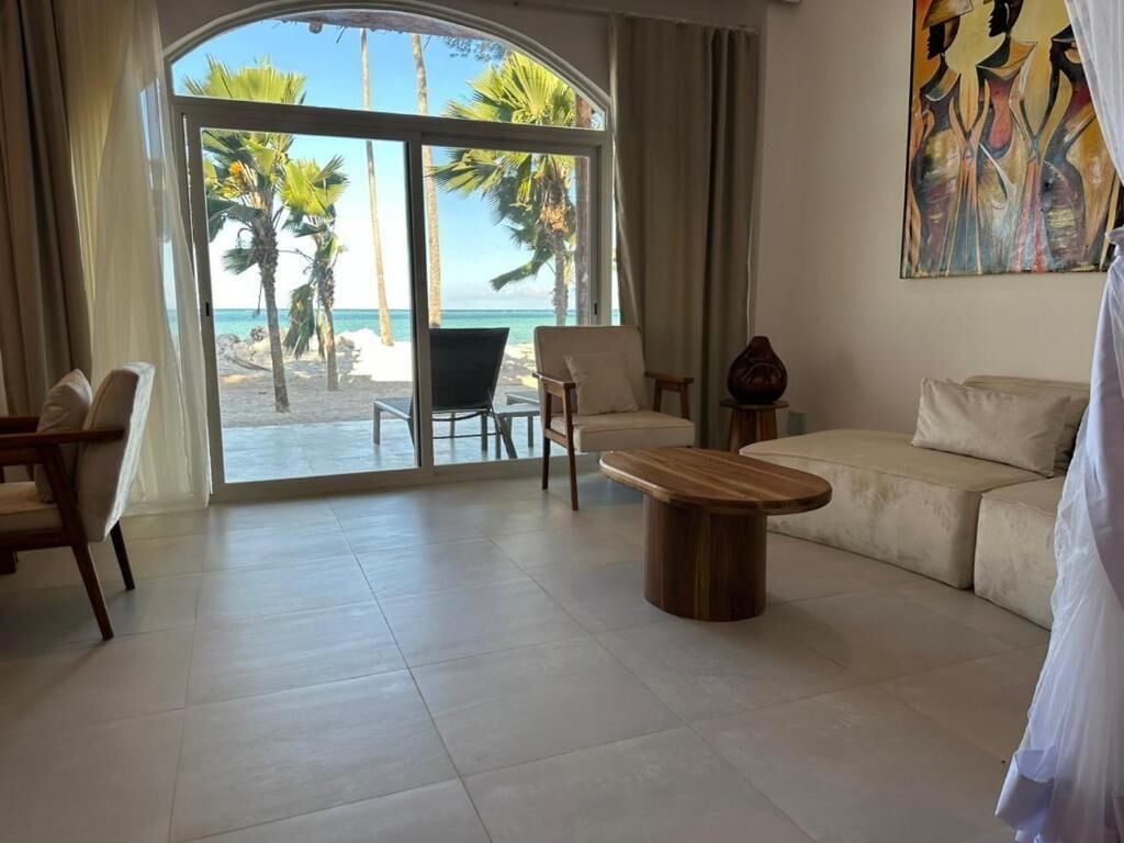 Amplia sala de estar con un gran ventanal corredizo que da a vista de playa, un sofá color beige, dos sillones y una mesa de centro de madera.