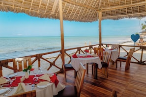 An outdoor dining area with tables set for two, overlooking a beach. - Royal Mandarin Hotel & Resort Zanzibar (Kiwengwa)