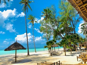 Een strand met rieten parasols, palmbomen en zand.