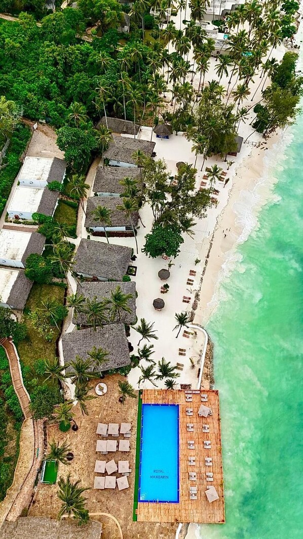 An aerial view of a resort with thatched-roof bungalows, a pool area, and a beach.
