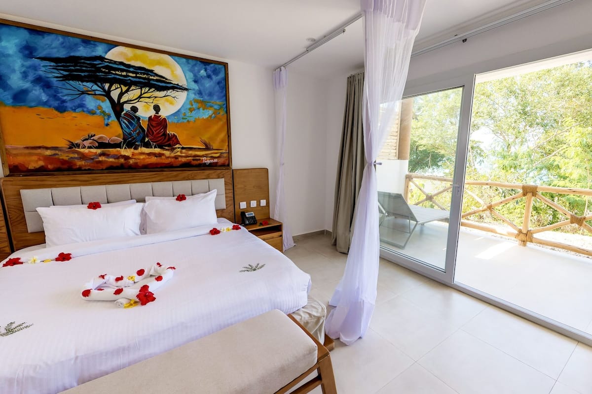 A hotel room with a large bed, a painting of a tree and two figures, a balcony with a view of greenery, and a window with sheer curtains.