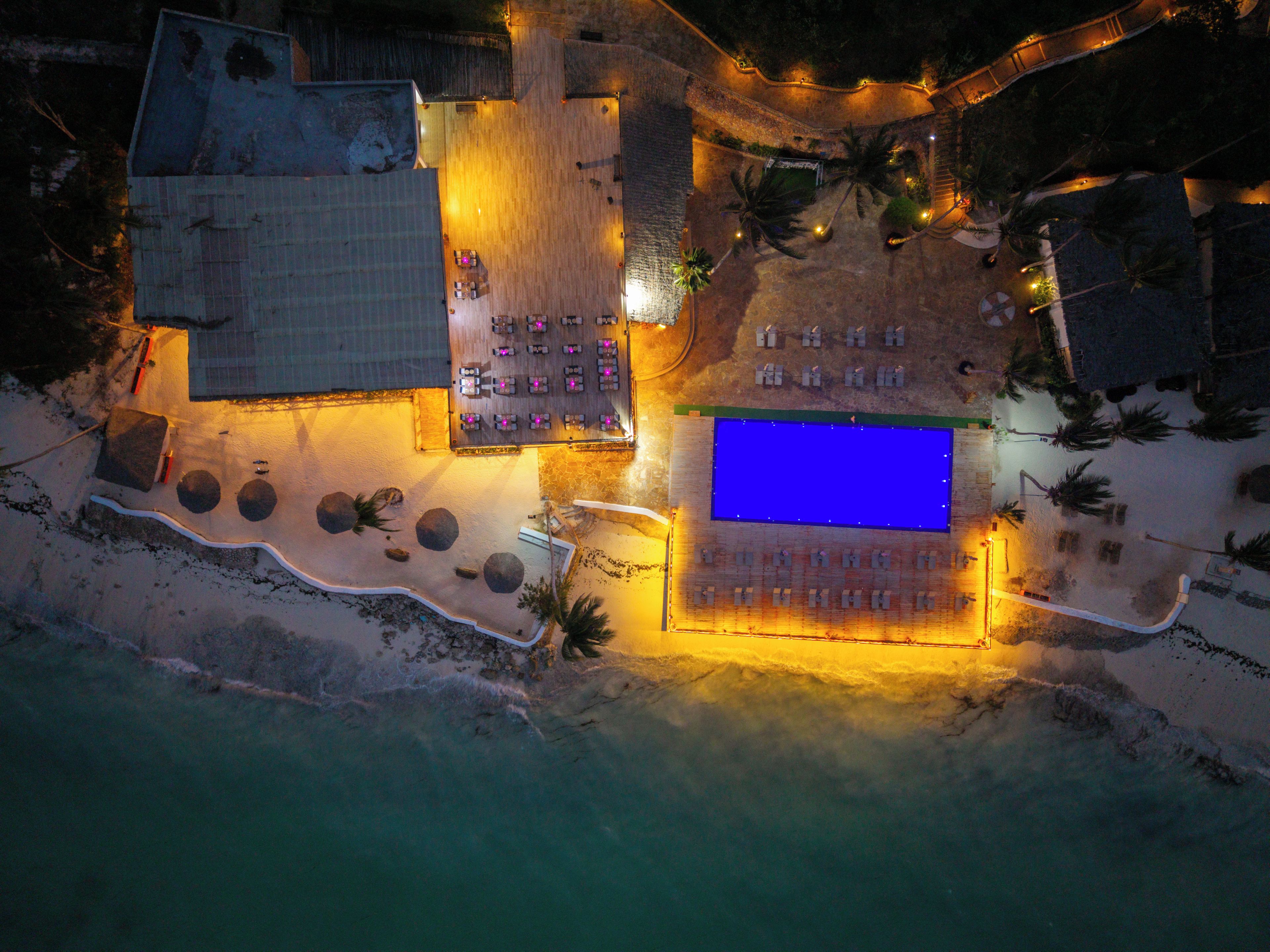 Vista aérea de un complejo turístico con piscina, zona de estar y asientos frente a la playa.