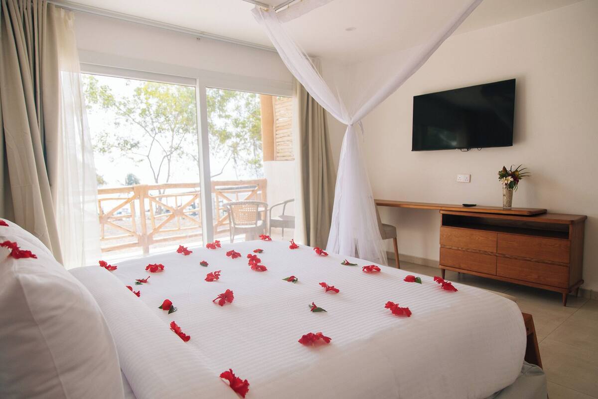 A hotel room with a bed adorned with red flowers, a television on the wall, and a balcony with wooden furniture.