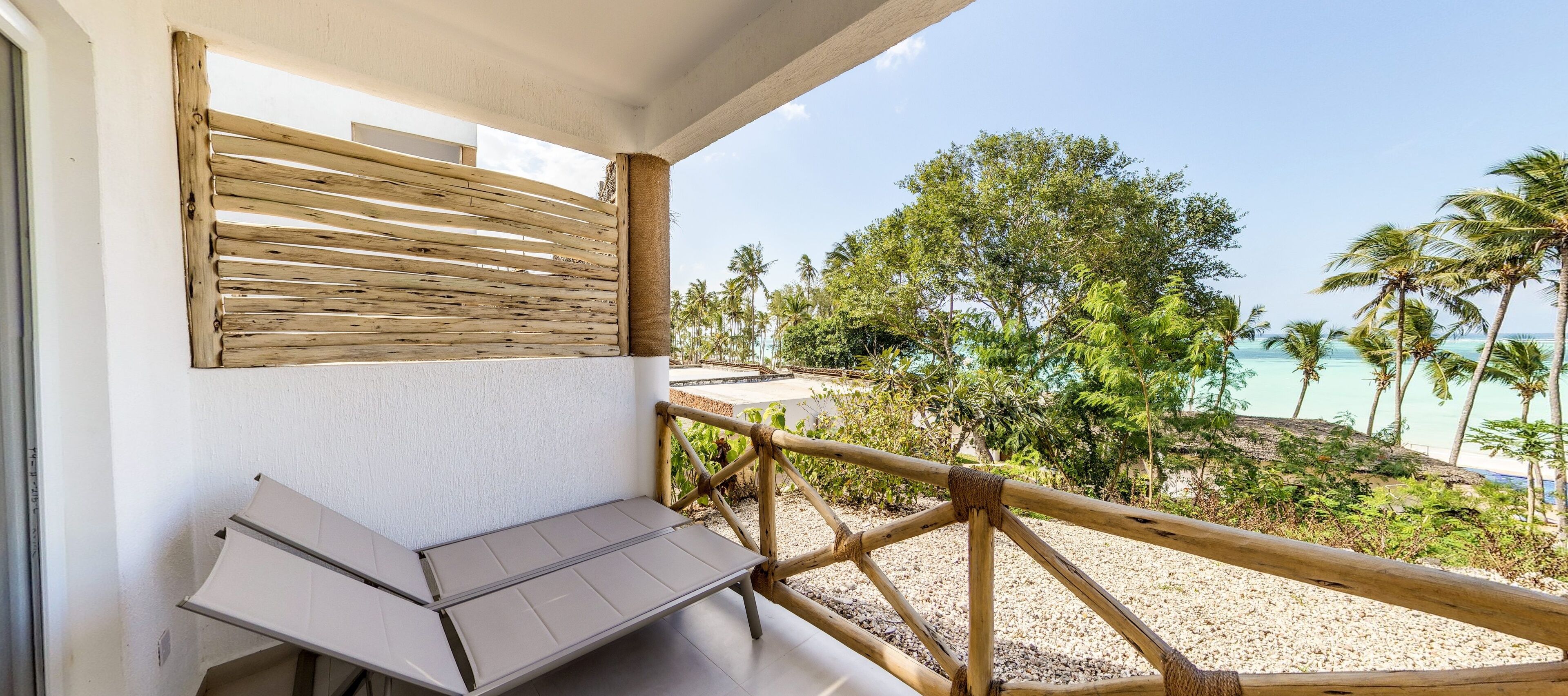 A balcony with a wooden railing, a white wall, and a view of the beach and palm trees.