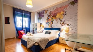 A bedroom with a large world map on the wall, a bed with a blue headboard, a red chair, and a desk with a lamp.