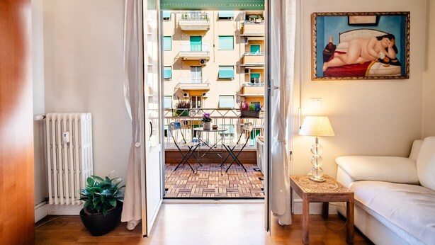 A hotel room with a wooden floor, a radiator, a potted plant, a small table with a lamp, a white sofa, and a balcony with a view of other buildings.