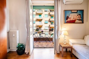 Una habitación de hotel con suelo de madera, un radiador, una planta en maceta, una mesita con lámpara, un sofá blanco y un balcón con vistas a otros edificios.