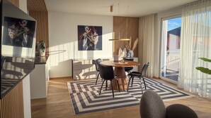Uma sala de estar moderna com uma mesa de jantar redonda, cadeiras pretas, piso de madeira e janelas amplas com cortinas leves.