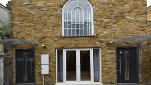 Un bâtiment en briques doté d’une grande fenêtre en arc de cercle, de deux portes noires et d’un petit espace extérieur surmonté d’un toit en métal.