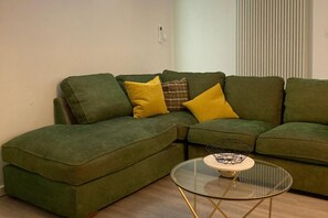 A green sectional sofa with yellow and plaid cushions, a glass coffee table with a metal base, and a window with vertical blinds.
