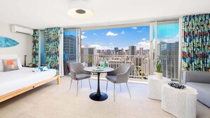 A modern hotel room with a bed, a round table with chairs, a sofa, and a large window offering a city view. - Waikiki studio w/ pool, sauna & city views (Honolulu)