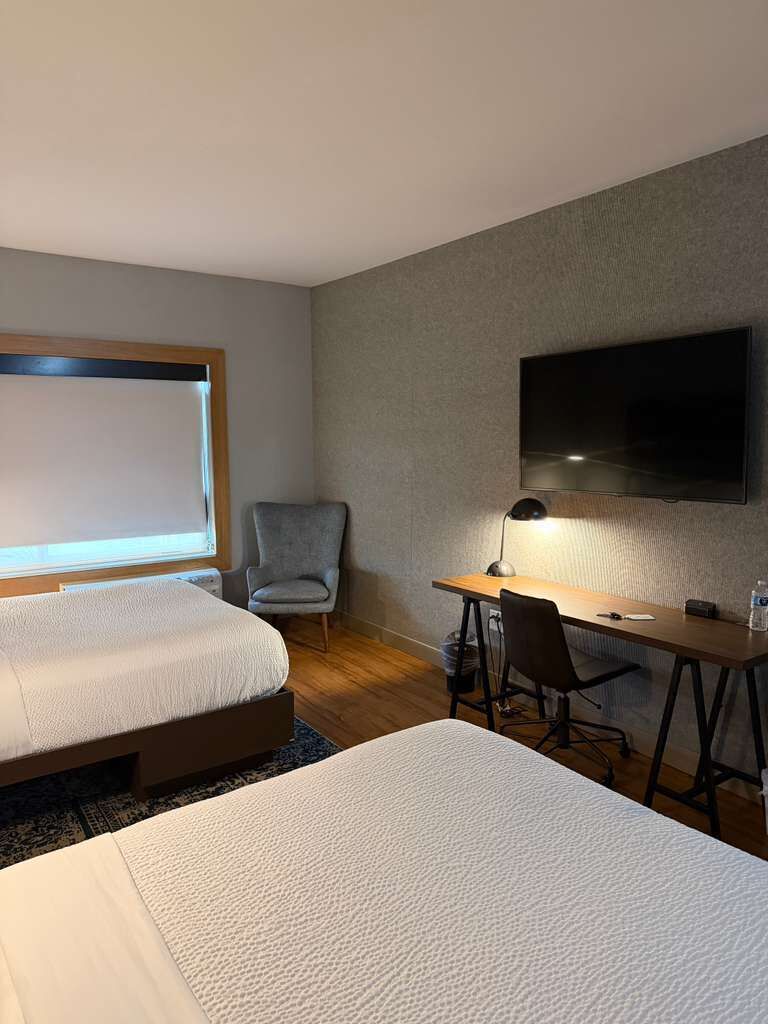 A hotel room with two beds, a desk, a chair, a TV, and a window.