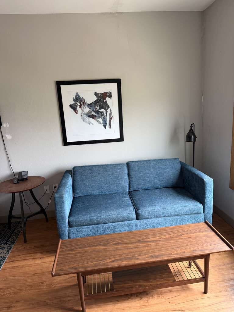 A living room with a blue sofa, a wooden coffee table, a framed abstract artwork on the wall, a small round table with a lamp, and a floor lamp.
