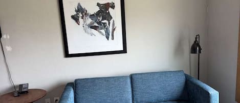 A living room with a blue sofa, a wooden coffee table, a framed abstract artwork on the wall, a small round table with a lamp, and a floor lamp.