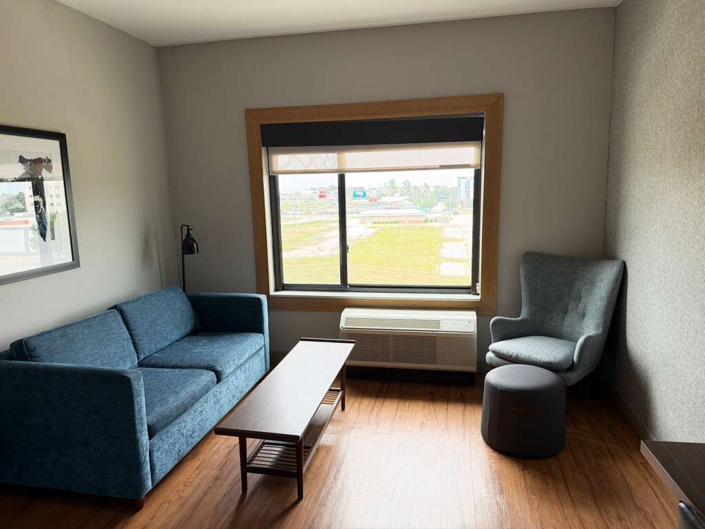 A room with a blue sofa, a grey armchair, a small ottoman, a wooden coffee table, and a large window with a view of a grassy area.