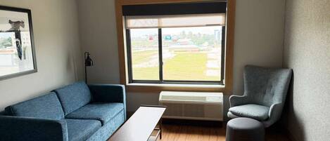 Una habitación con un sofá azul, un sillón gris, un pequeño reposapiés, una mesa de centro de madera y un ventanal con vistas a un área verde.