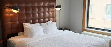 A bedroom with a large bed, a headboard, bedside lamps, a window, and a wall with framed pictures.