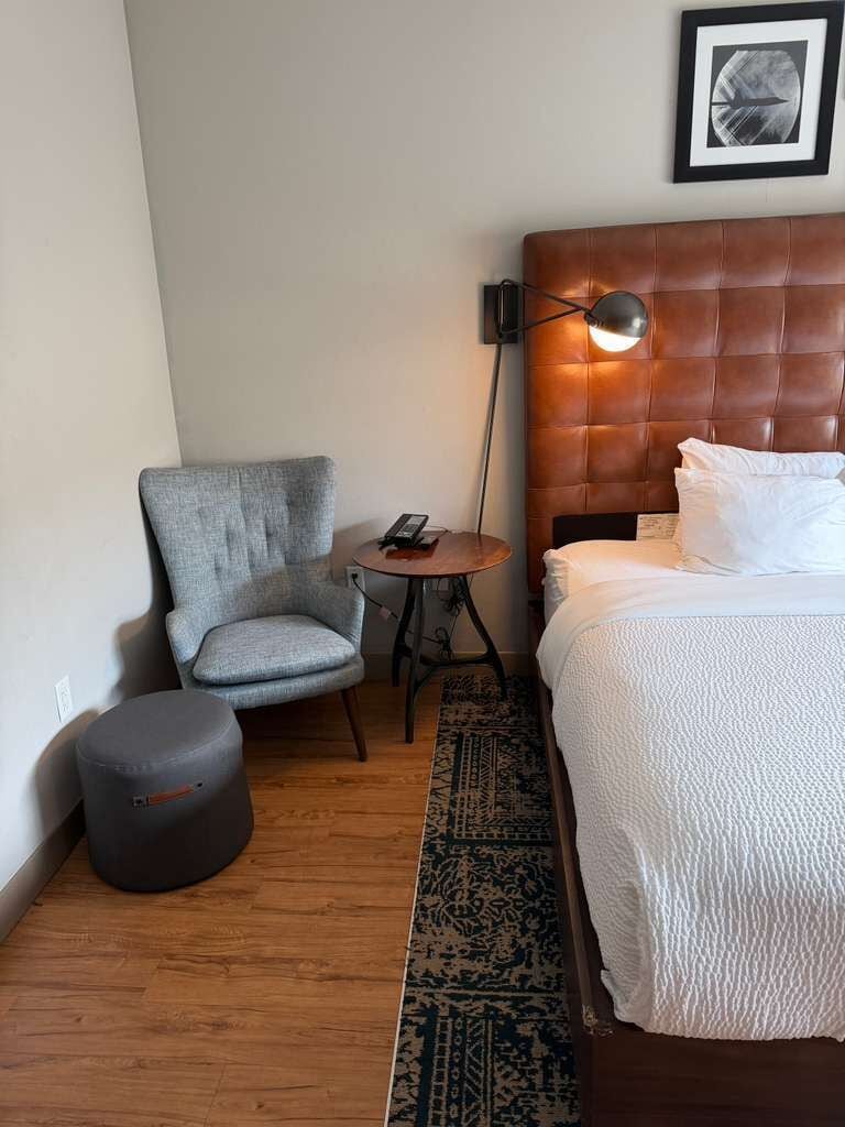 Una habitación de hotel con cama, una silla, una mesita, una lámpara y una alfombra.
