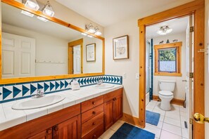 Une salle de bain avec deux lavabos, des meubles en bois, un grand miroir et une fenêtre donnant sur les arbres.