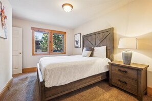 Une chambre avec un grand lit, une tête de lit en bois, une table de chevet avec une lampe et une fenêtre donnant sur des arbres.