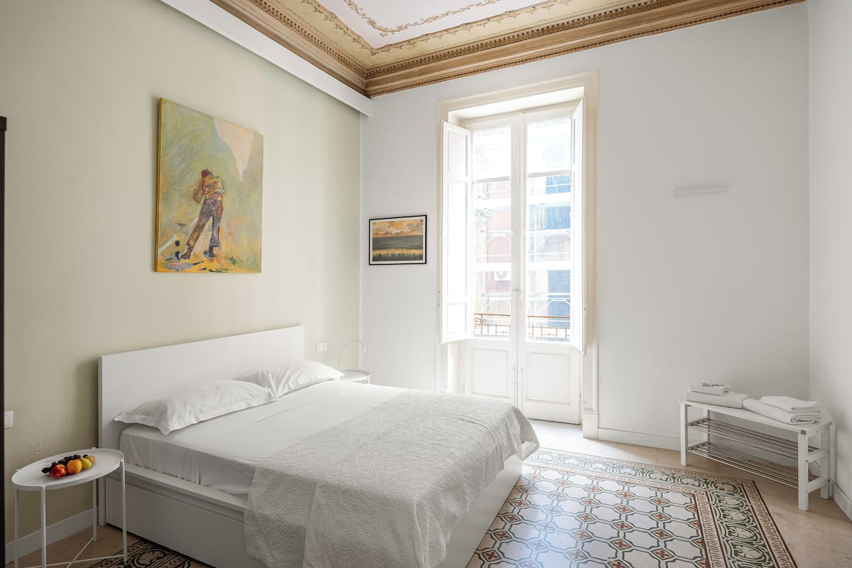 A bedroom with a bed, a small table with fruit, a painting, and a view through a window.
