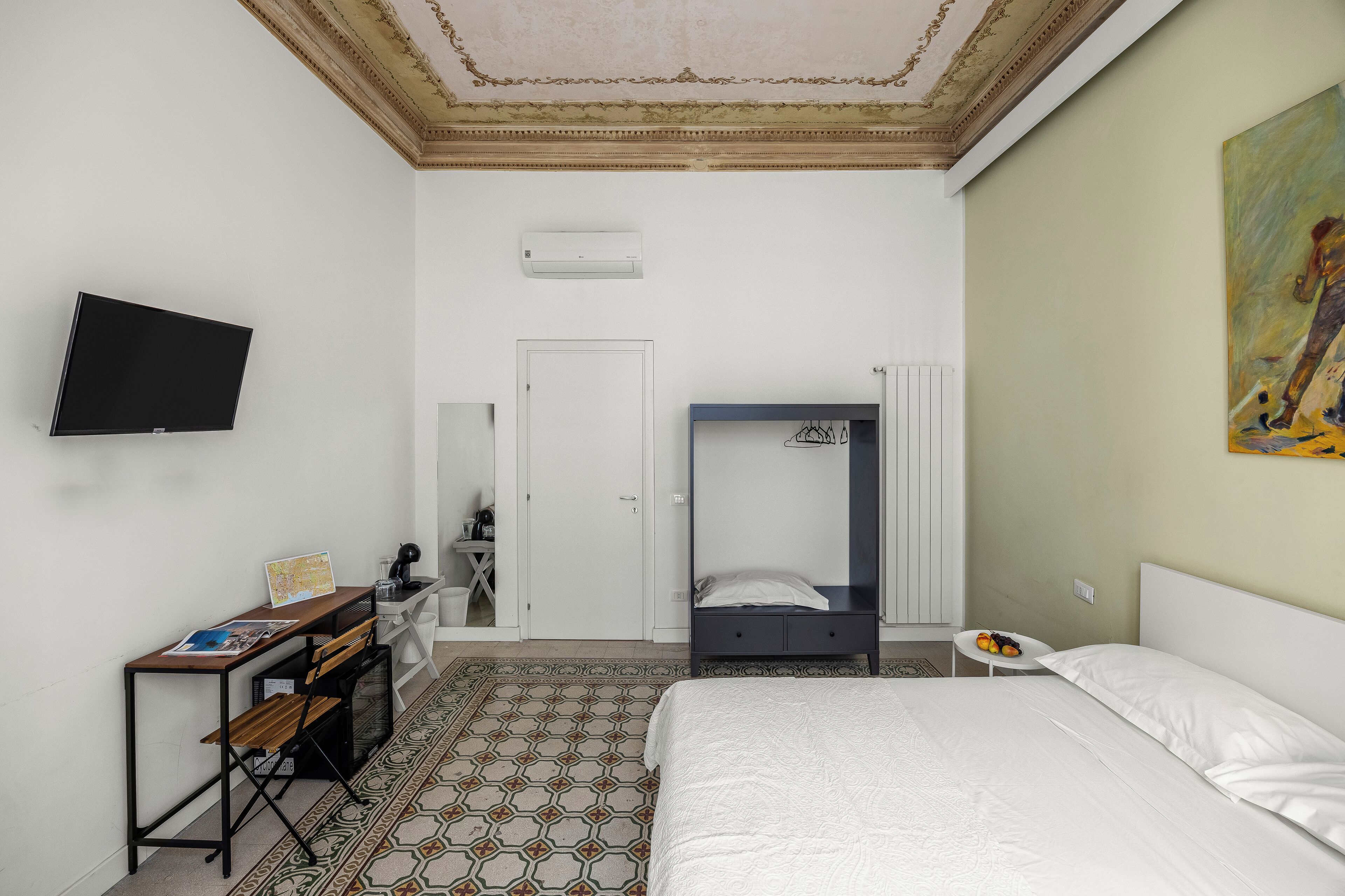 A hotel room with a bed, a desk with a chair, a television mounted on the wall, and a painting on the wall.