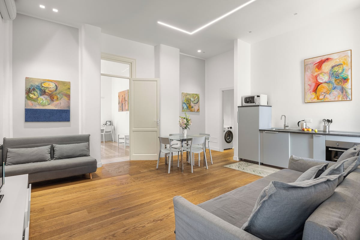 A modern living room with a grey sofa, a dining area with a table and chairs, and a kitchen with a microwave and sink.
